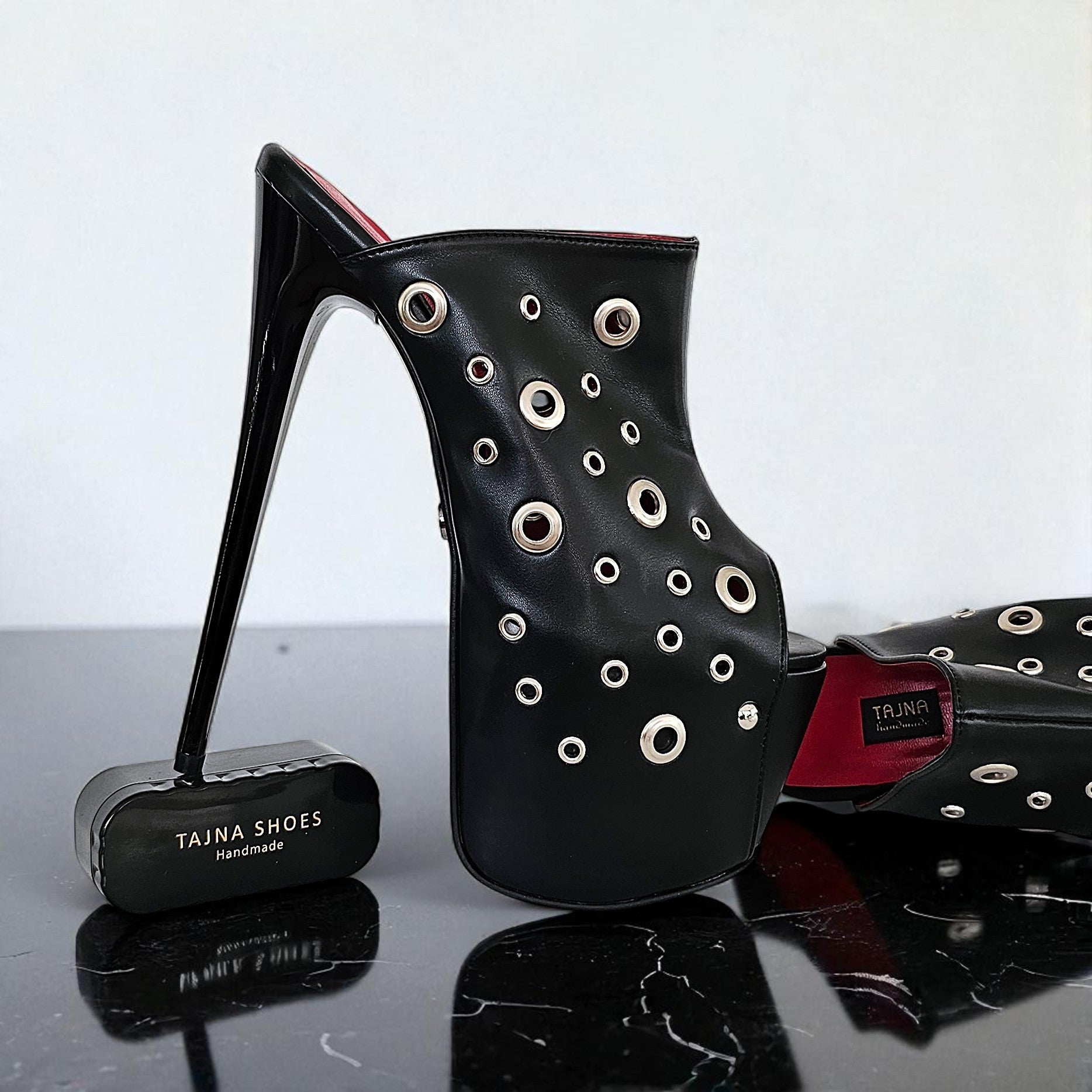 Black high-heeled shoe with eyelet details on a reflective surface, branded 'Tajna Shoes'.