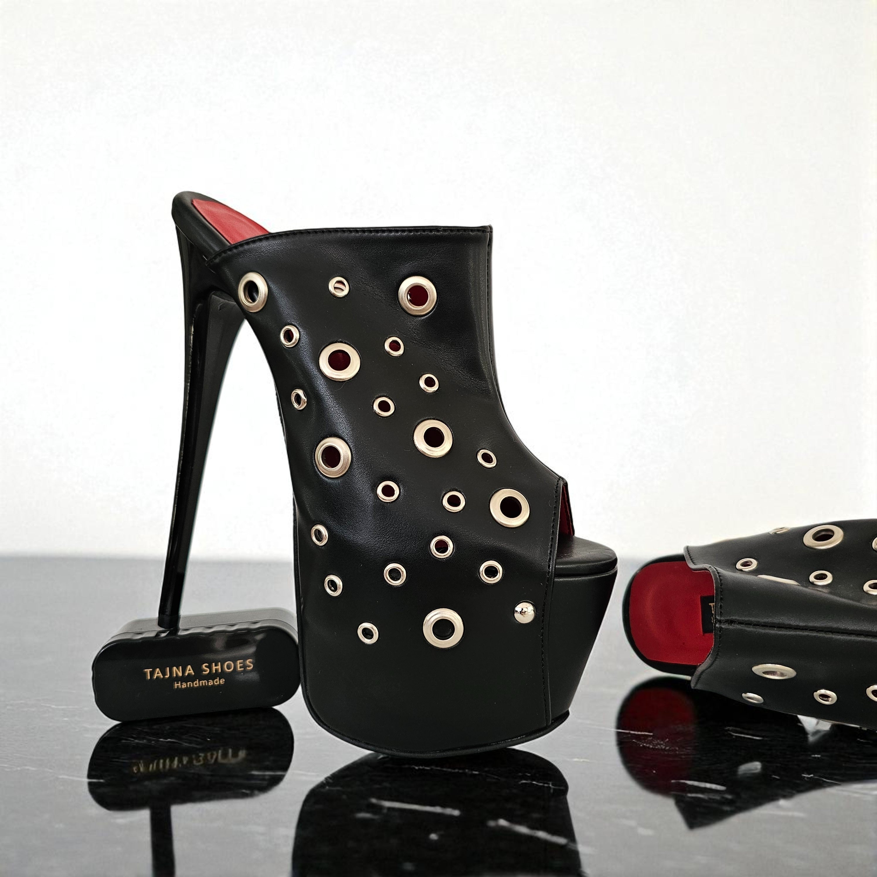 Black high-heeled shoes with circular cutouts on a reflective surface, featuring the brand 'Tajna Shoes'.