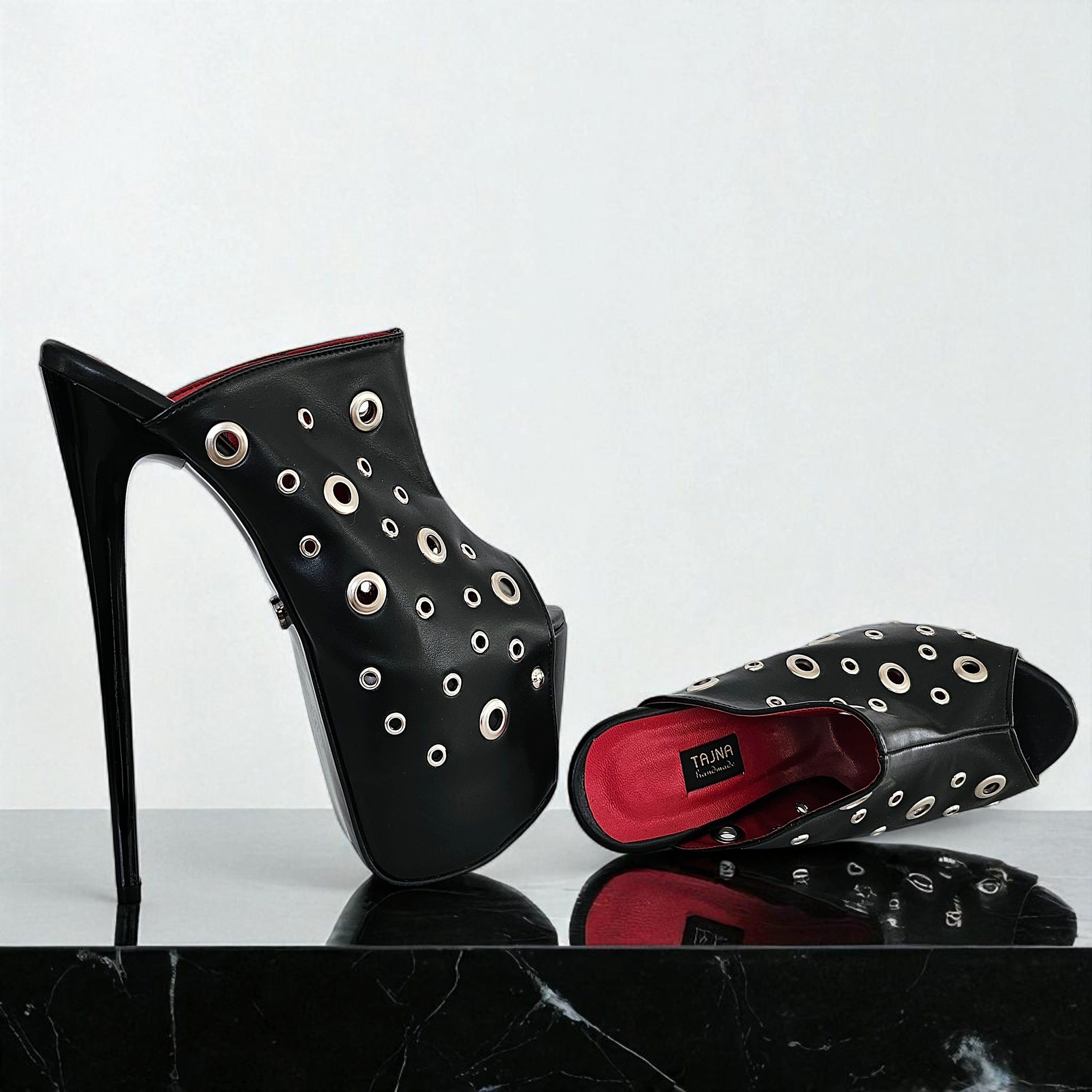 Black high-heeled shoes with grommet details on a reflective surface.