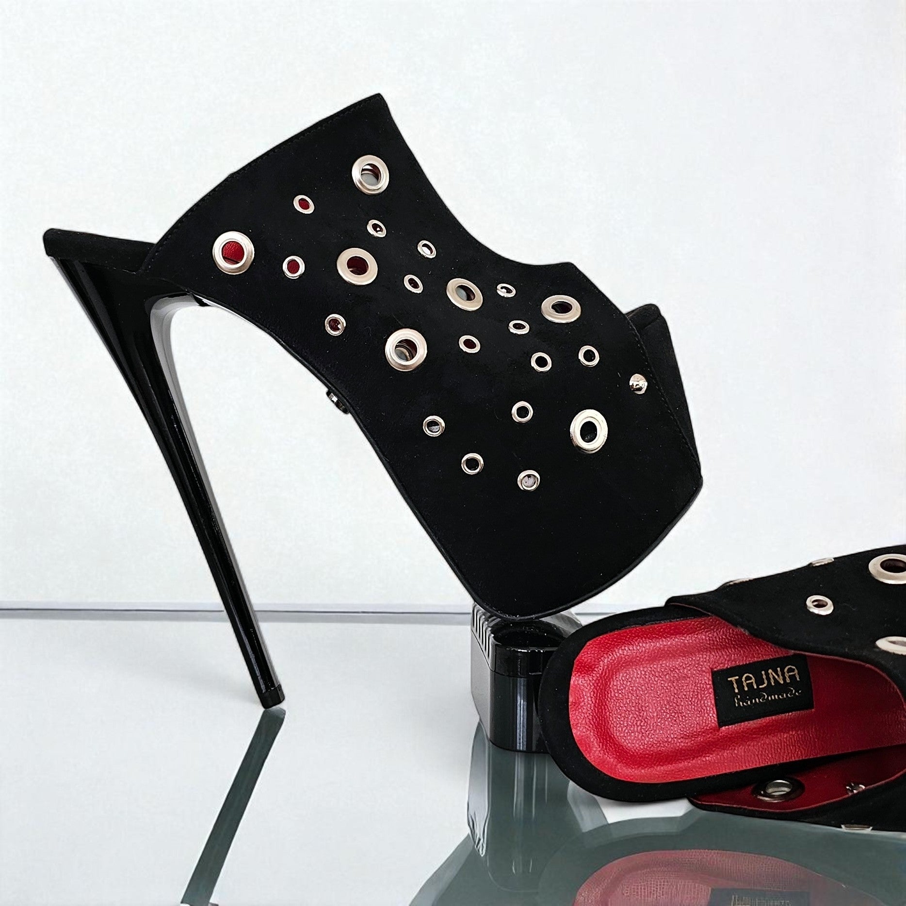 Black high-heeled shoe with eyelets and red insole on a reflective surface